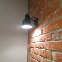 Kinkiet w stylu industrialnym, zamontowany na ścianie z czerwonej cegły, oświetlający gładką, szarą ścianę.