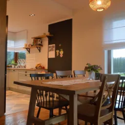 Jasna kuchnia z drewnianym stołem i krzesłami, dekoracyjnymi lampami wiszącymi z rattanu oraz czarną tablicą na ścianie.