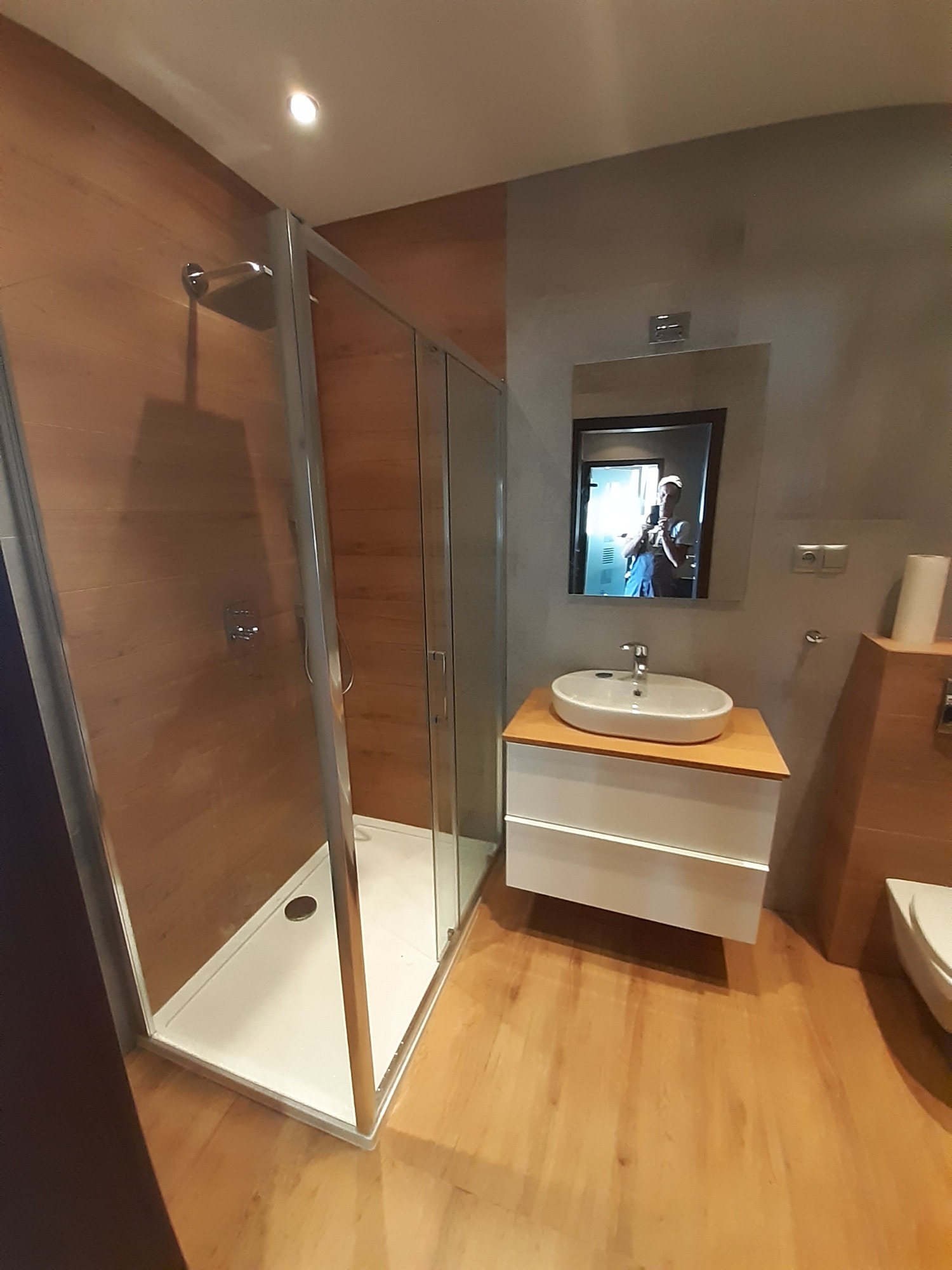 Nowoczesna łazienka z kabiną prysznicową typu walk-in, umywalką nablatową na szafce z drewnianym blatem i toaletą; widoczne odbicie osoby robiącej zdjęcie w lustrze.
