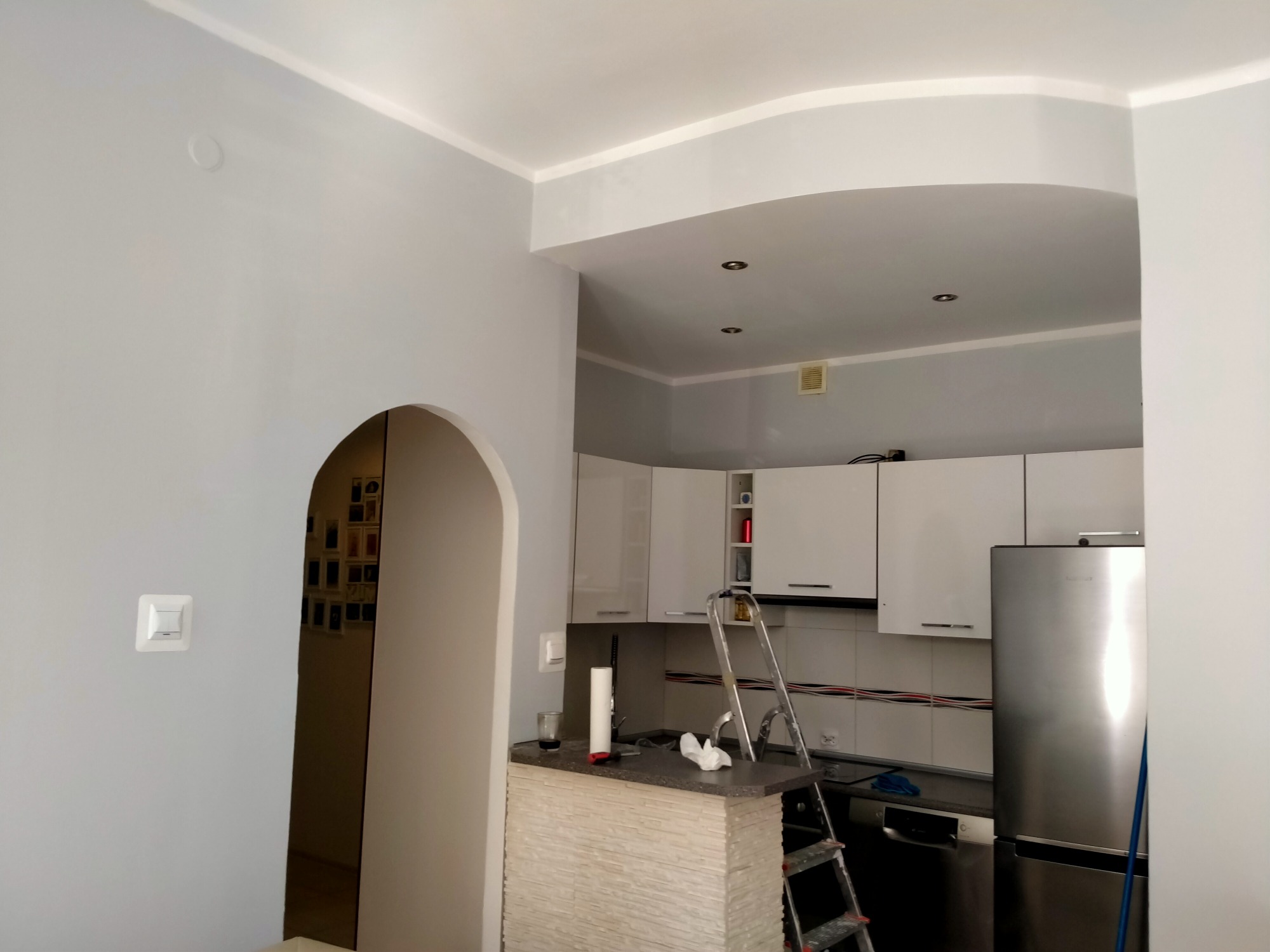 Wnętrze kawalerki w trakcie malowania: szare ściany, białe meble kuchenne, aluminiowa drabina, widoczny fragment łuku drzwiowego.