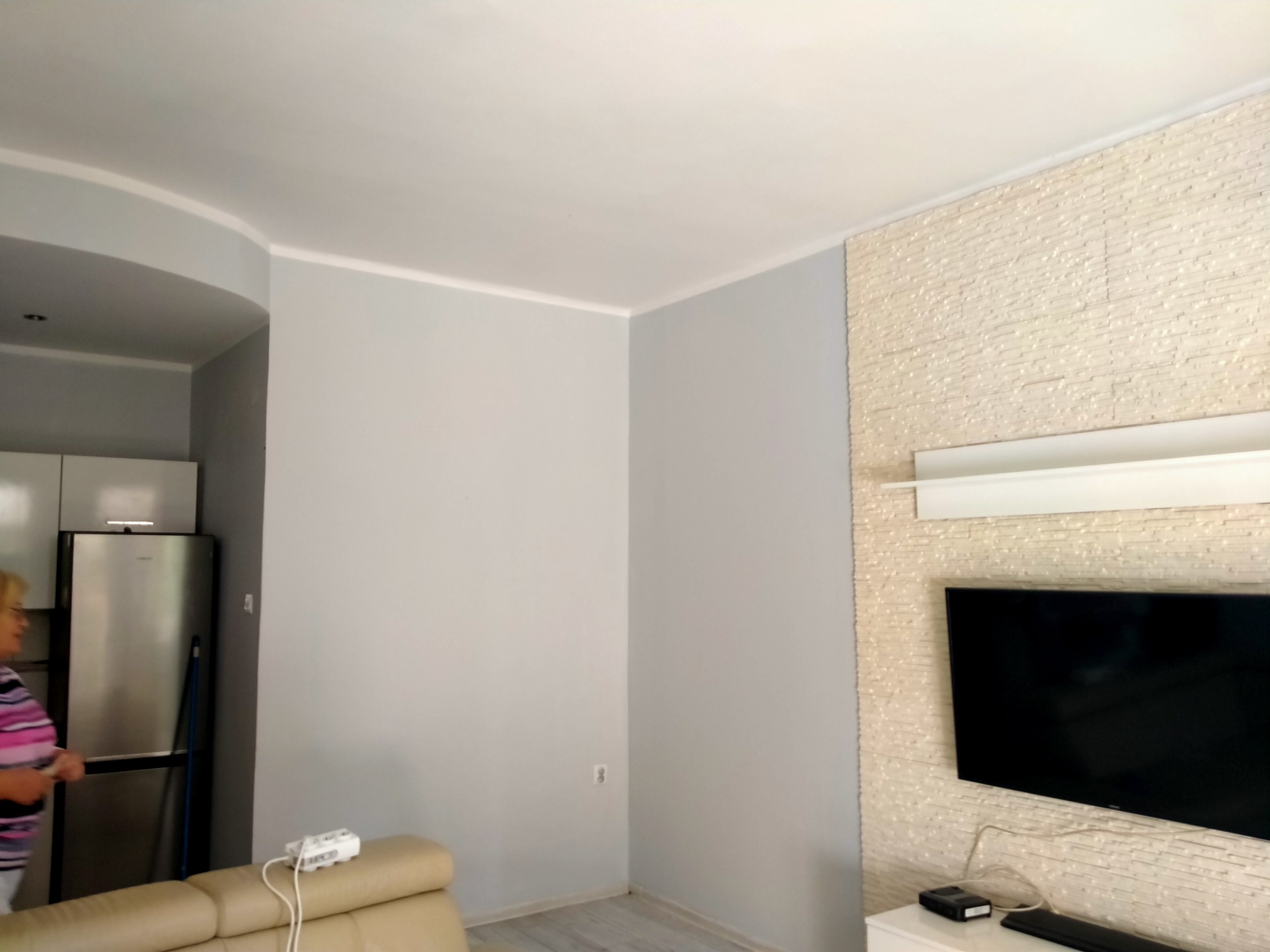 Jasnoszary salon z dekoracyjną ścianą z jasnego kamienia, na której wisi telewizor. Widoczny fragment beżowej sofy i osoba w tle.