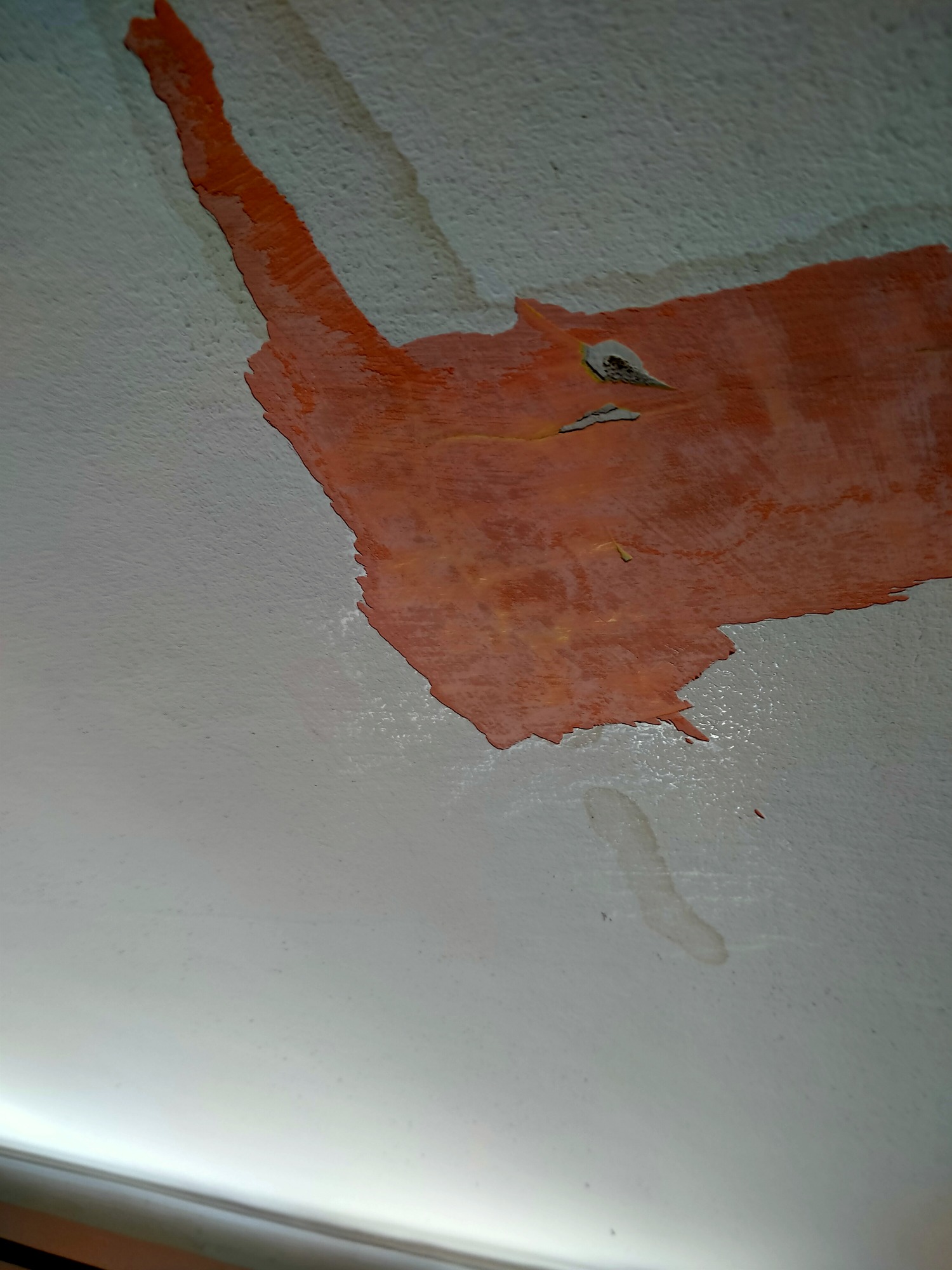 Fragment ściany z odpryskującą, pomarańczową farbą, ukazujący warstwę podkładową i drobne uszkodzenia powierzchni.