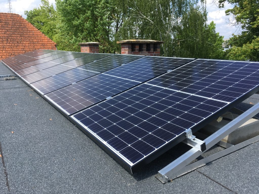 Monokrystaliczne panele na płaskim dachu z szarym pokryciem, zamocowane na srebrnym stelażu, widoczne kominy i drzewa w tle.