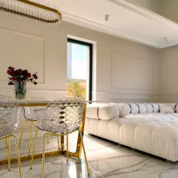 Eleganckie wnętrze w odcieniach bieli i złota: marmurowa podłoga, transparentne krzesła z pozłacanymi nogami, designerska lampa nad stołem z bukietem czerwonych róż, w tle modułowa sofa i okno...