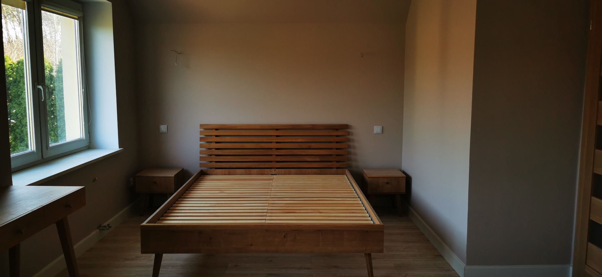 Surowa drewniana rama łóżka z listewkowym zagłówkiem i pasującymi szafkami nocnymi w pokoju z beżowymi ścianami, widoczne niedokończone prace elektryczne.