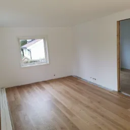 Jasne panele podłogowe z widocznym wzorem drewna w nowo wykończonym pokoju z białymi ścianami i oknem wychodzącym na zewnątrz. Listwy przypodłogowe leżą obok ściany.