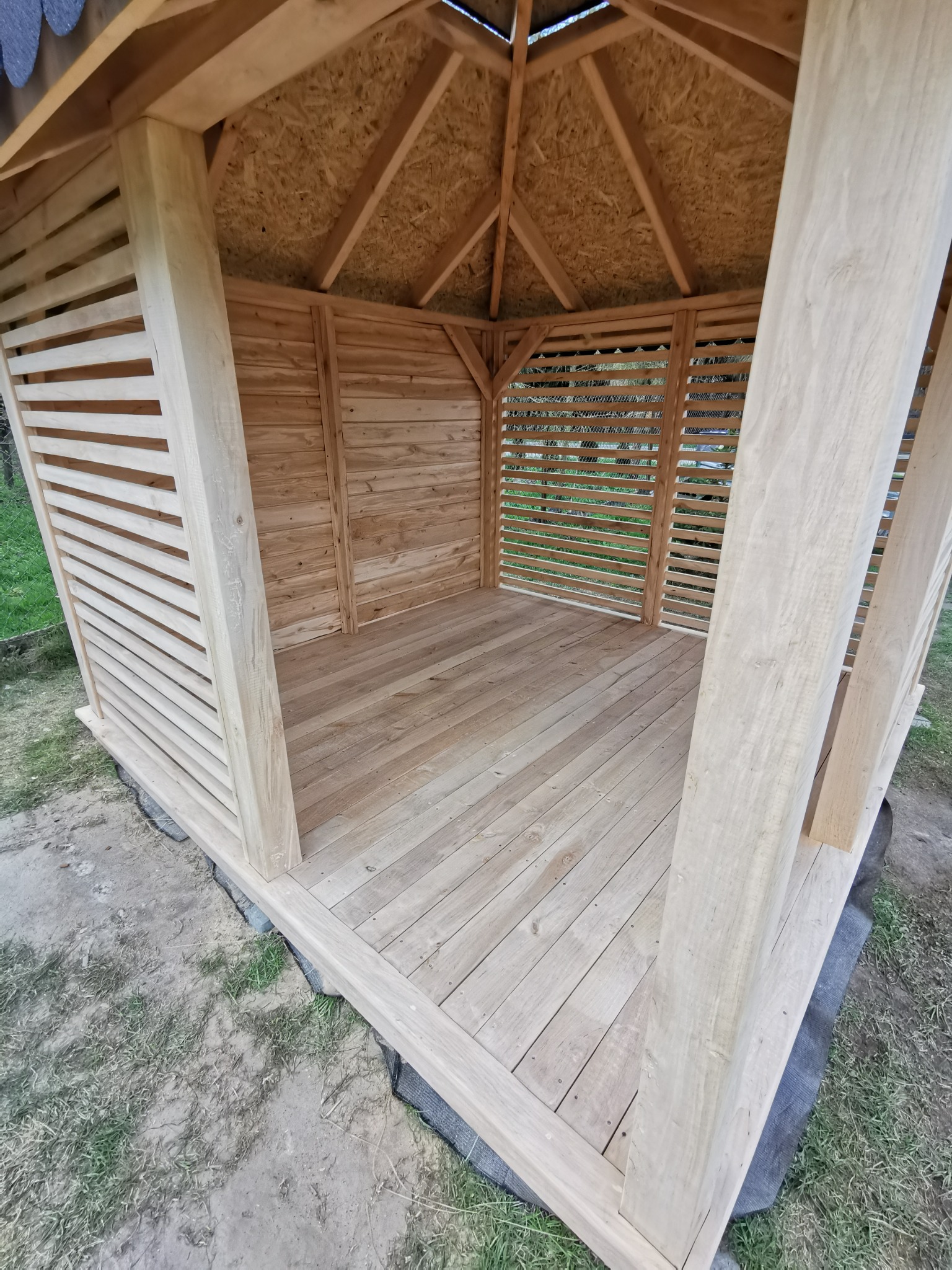 Surowa drewniana altana z lamelowymi ścianami i drewnianą podłogą, widok od wewnątrz, świeżo po montażu, na zewnątrz widoczna trawa i ziemia.