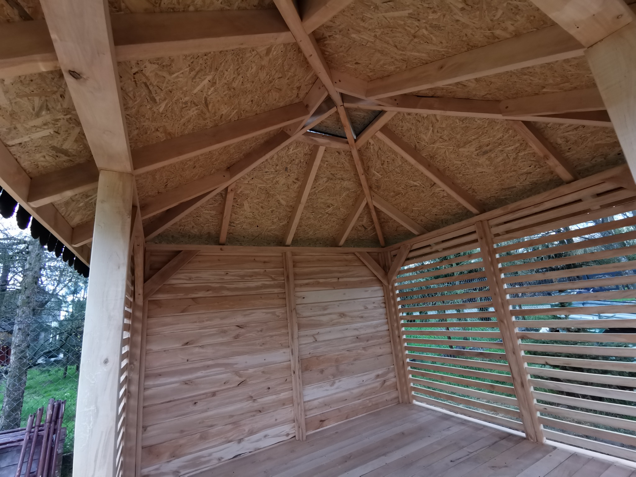 Wnętrze drewnianej altany z widoczną konstrukcją dachu z desek i płyt OSB, drewniana podłoga i ażurowe ściany boczne, surowe drewno przed impregnacją.