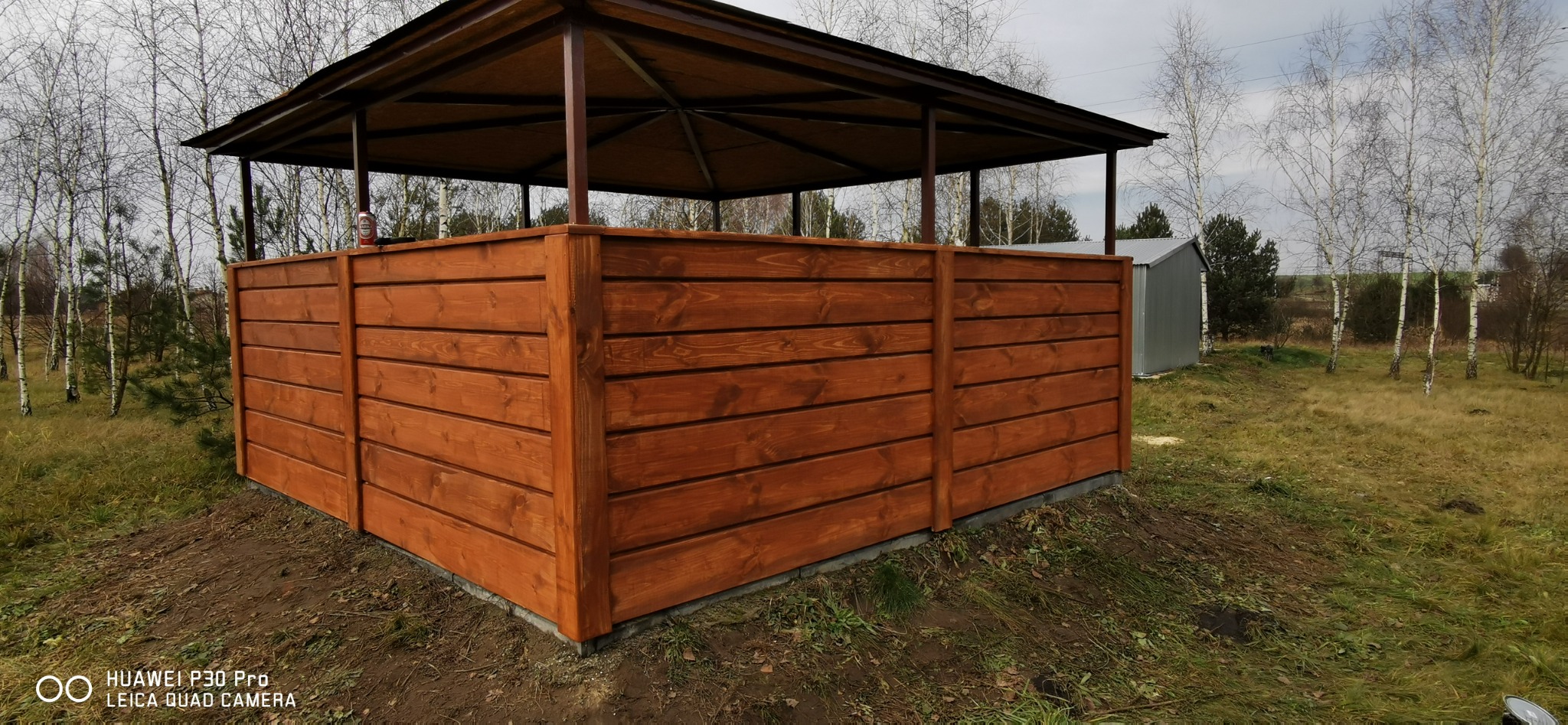 Drewniana altana ogrodowa z brązowymi panelami i ciemnym dachem na tle jesiennego krajobrazu z brzozami i metalowym garażem.