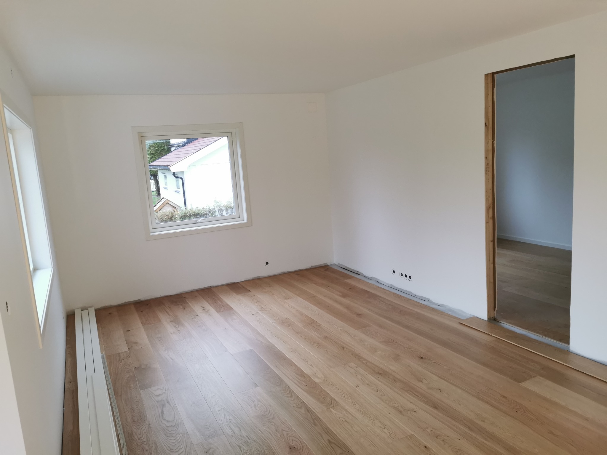 Jasne panele podłogowe z widocznym wzorem drewna w nowo wykończonym pokoju z białymi ścianami i oknem wychodzącym na zewnątrz. Listwy przypodłogowe leżą obok ściany.