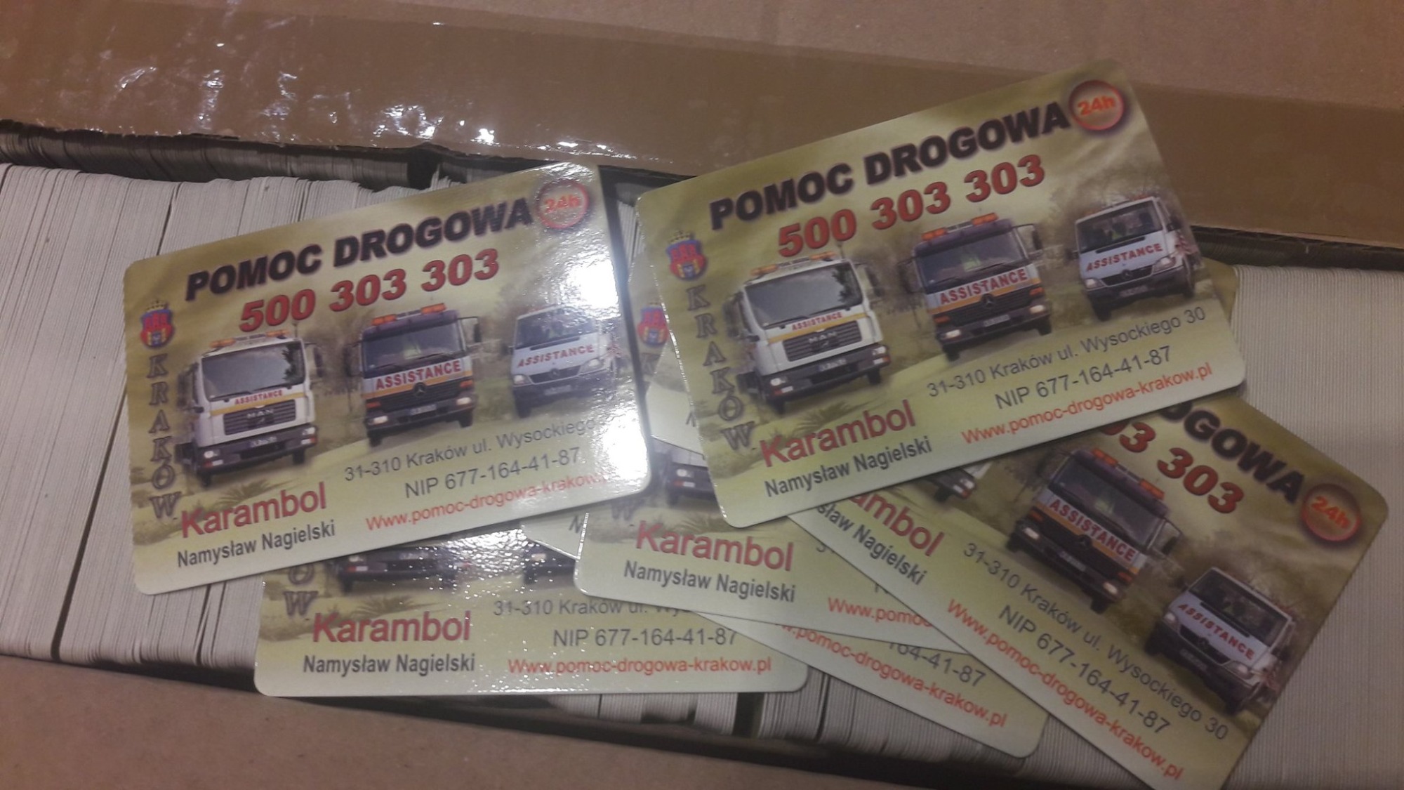 Stos wizytówek firmy pomocy drogowej Karambol z Krakowa, prezentujących numery telefonów i logotypy pojazdów assistance na jasnym tle.