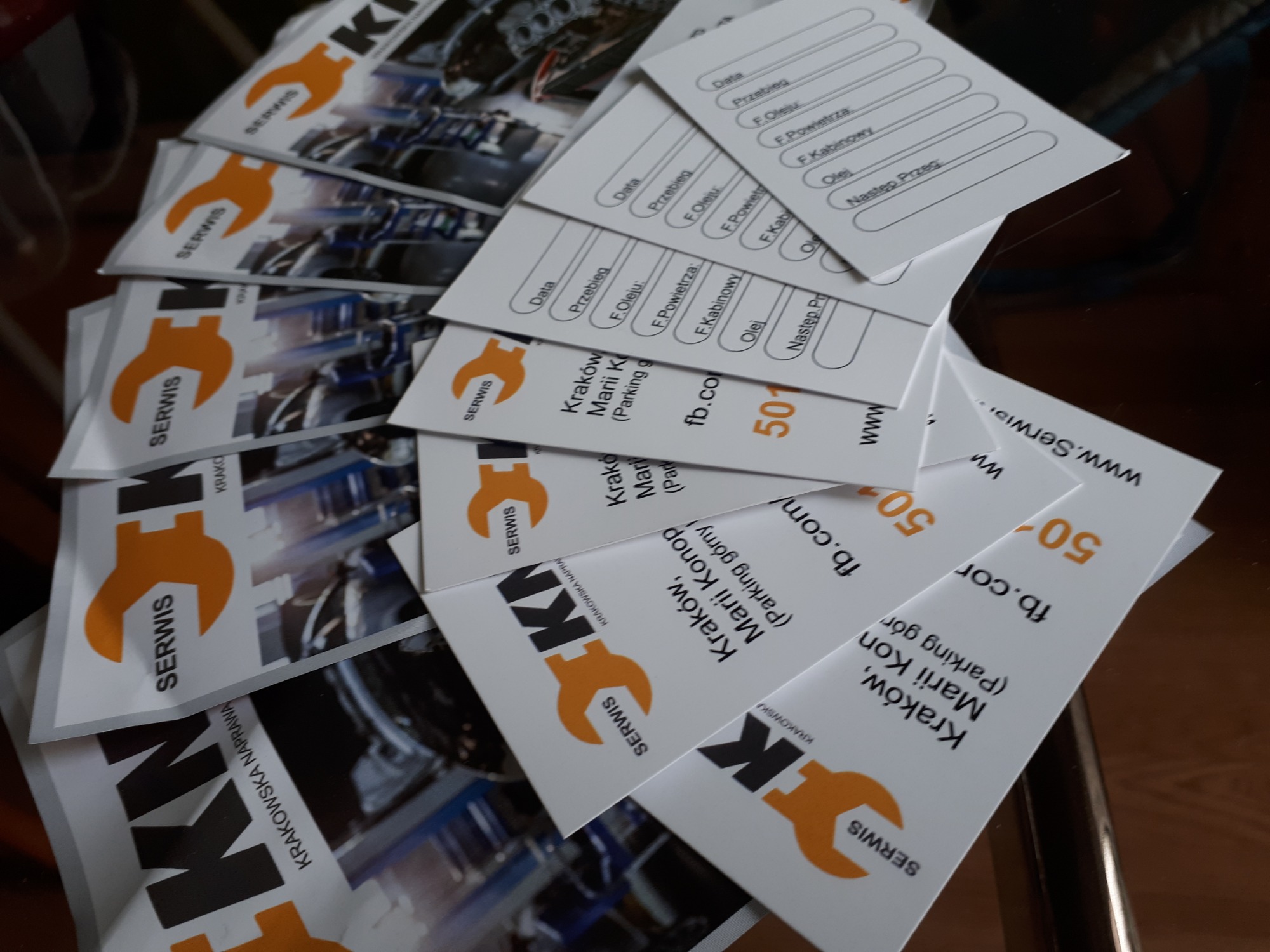 Stos wizytówek serwisu samochodowego z pomarańczowym kluczem francuskim i danymi kontaktowymi firmy z Krakowa oraz formularzem do wpisywania daty, przebiegu i wymiany filtrów i oleju.