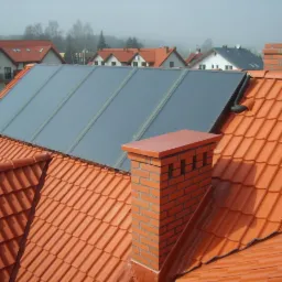 Kolektory słoneczne na dachu z czerwonej dachówki i ceglanym kominem. W tle zabudowa jednorodzinna w lekkiej mgle.