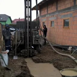 Wiercenie studni głębinowej przy budynku w trakcie budowy. Dwóch mężczyzn obsługuje wiertnicę. Na pierwszym planie czarny płot i akcesoria do wiercenia.