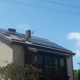 Dom jednorodzinny z zamontowanymi panelami fotowoltaicznymi na dachu, widok z dołu na tle błękitnego nieba z pojedynczymi chmurami.