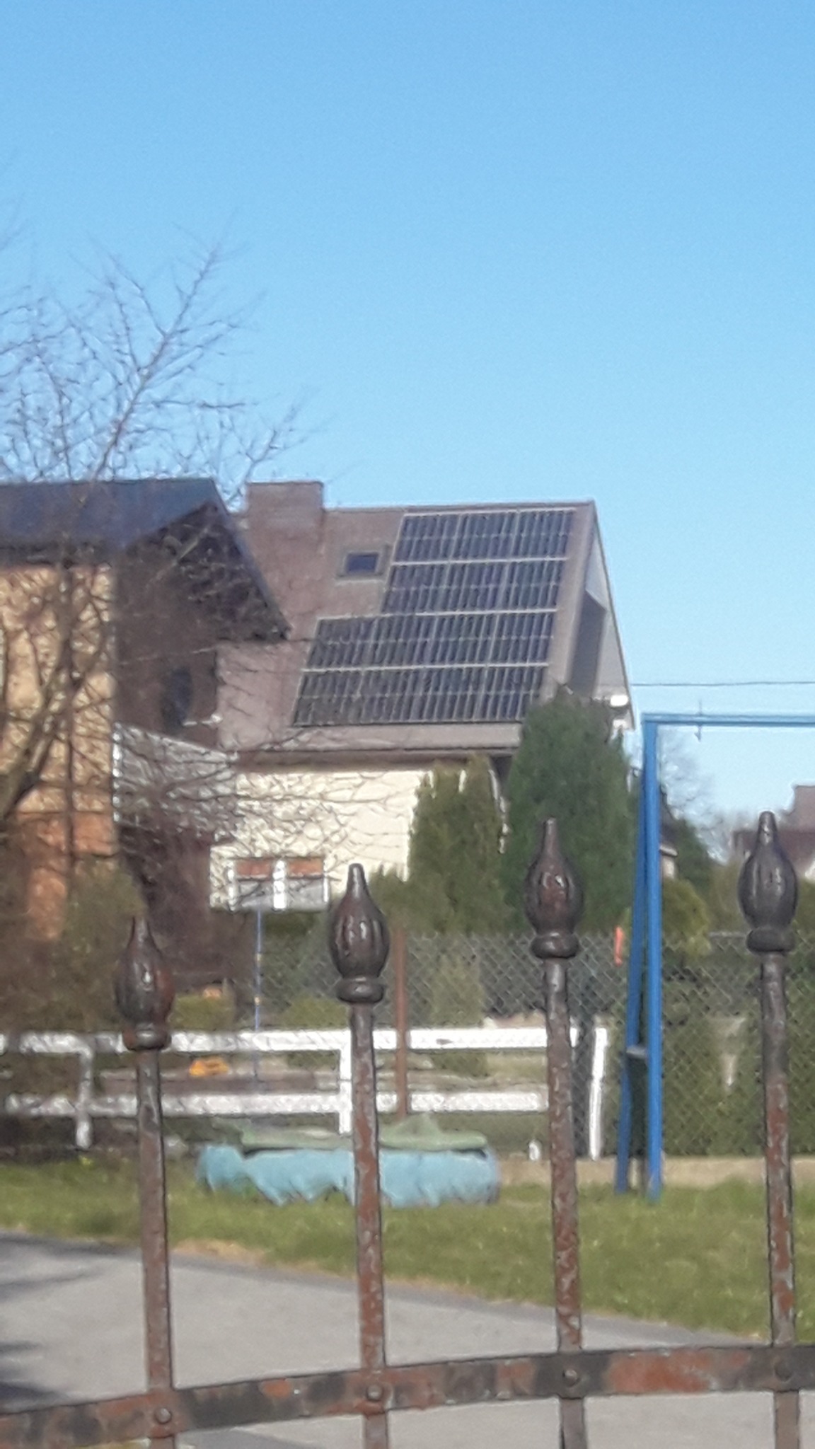 Panele słoneczne na dachu domu, widoczne zza metalowego ogrodzenia z ozdobnymi zakończeniami, w tle drzewa i błękitne niebo.