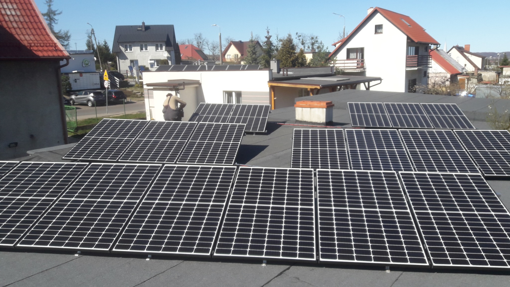 Montaż paneli fotowoltaicznych na dachu domu jednorodzinnego; widoczny instalator w kombinezonie, panele ułożone w rzędach, w tle zabudowa mieszkalna i błękitne niebo.