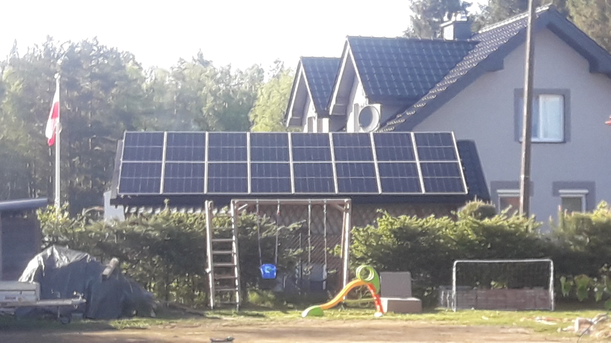 Panele słoneczne na dachu budynku gospodarczego, w tle dom jednorodzinny z anteną satelitarną i powiewająca flaga Polski, na pierwszym planie plac zabaw.