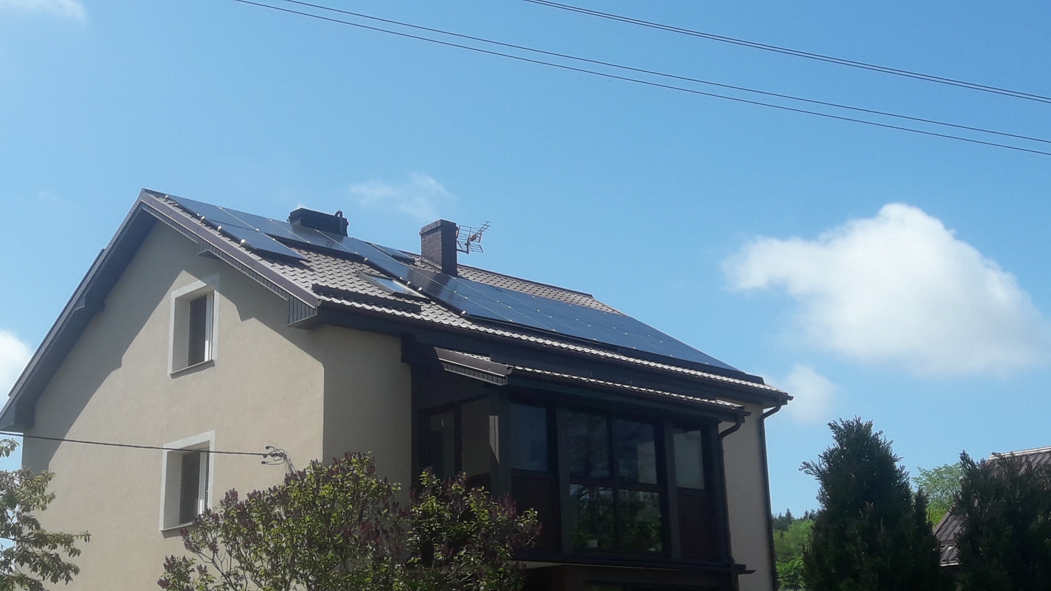 Dom jednorodzinny z zamontowanymi panelami fotowoltaicznymi na dachu, widok z dołu na tle błękitnego nieba z pojedynczymi chmurami.