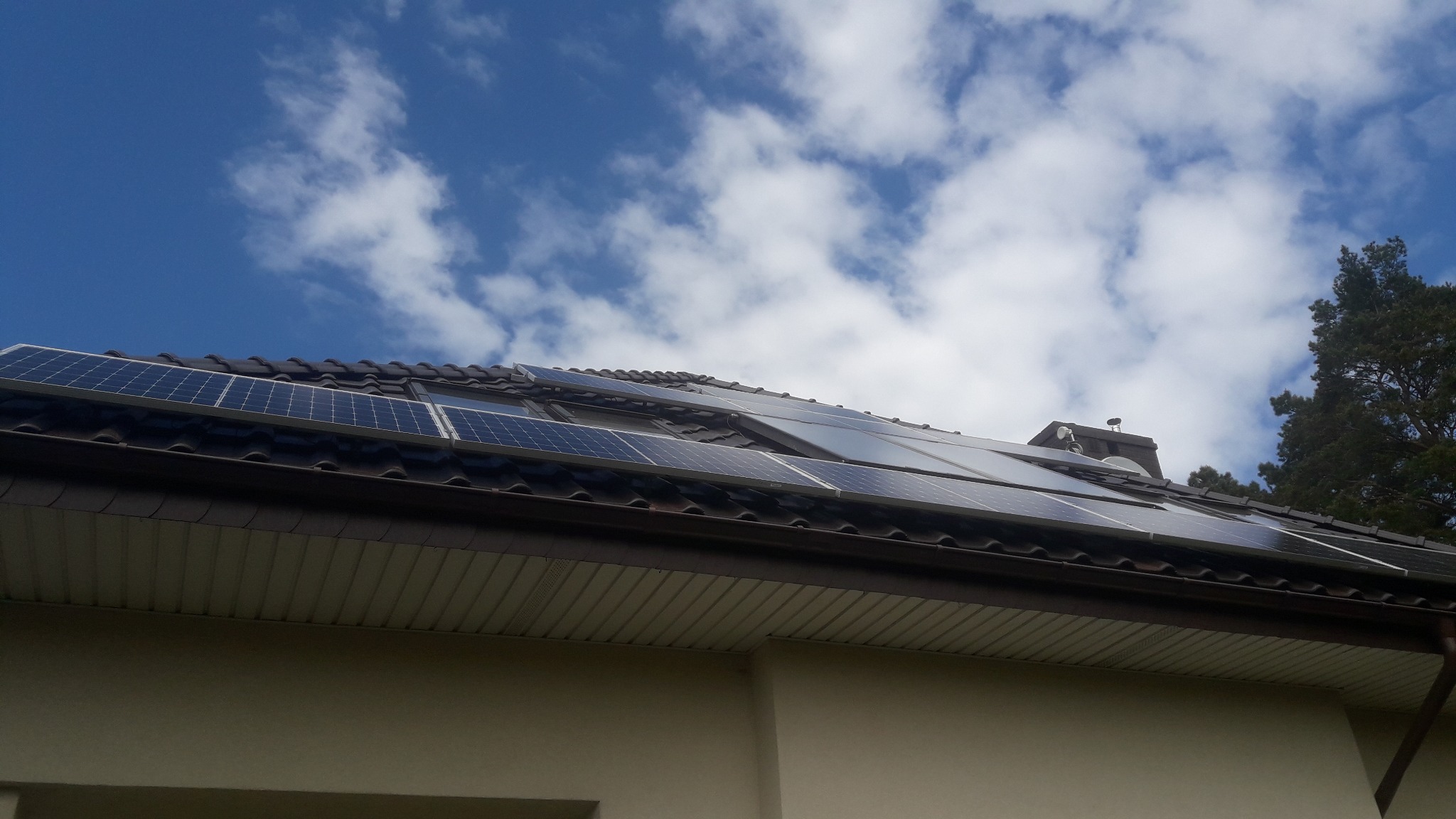 Panele fotowoltaiczne zamontowane na brązowym dachu domu jednorodzinnego, widok z dołu na tle błękitnego nieba z chmurami.