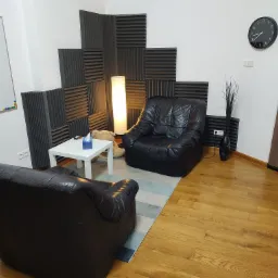 Pokój z dwoma czarnymi skórzanymi fotelami, białym stolikiem z chusteczkami, lampą podłogową i panelami akustycznymi na ścianie, tworzący spokojną przestrzeń.