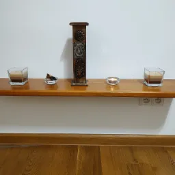 Drewniana półka na białej ścianie z dwoma świecami w szklanych pojemnikach, drewnianą ozdobą z metalowymi wstawkami i małą szklaną miseczką z kawałkami drewna, oświetlona świecą wewnątrz,...