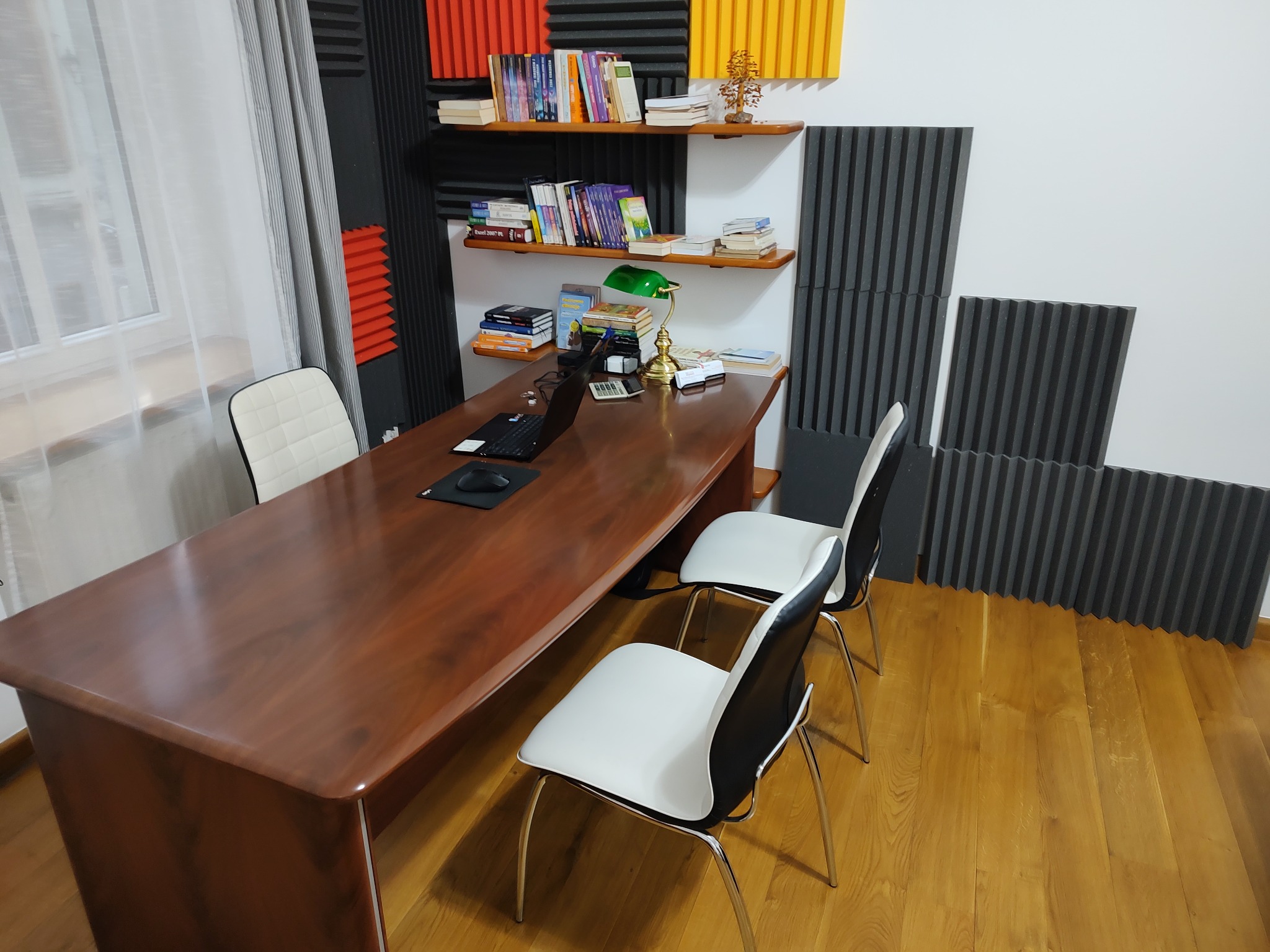 Gabinet z drewnianym biurkiem, laptopem, lampką, książkami i panelami akustycznymi na ścianach.