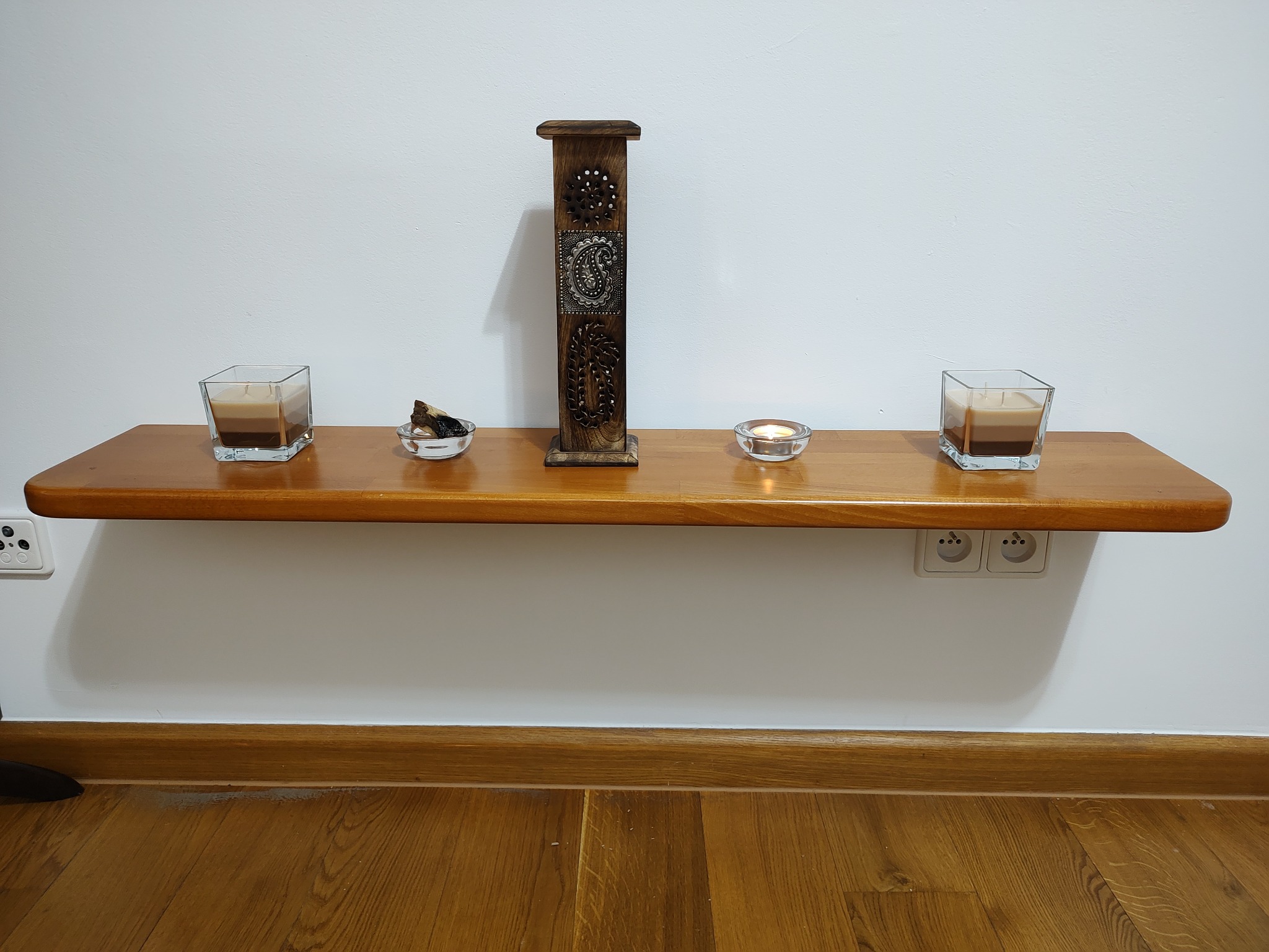 Drewniana półka na białej ścianie z dwoma świecami w szklanych pojemnikach, drewnianą ozdobą z metalowymi wstawkami i małą szklaną miseczką z kawałkami drewna, oświetlona świecą wewnątrz,...