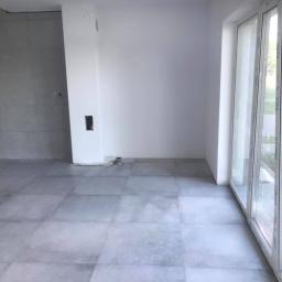 TopHome Usługi remontowo - budowlane - Wnętrze pokoju po remoncie z szarymi płytkami na podłodze i świeżo pomalowanymi na biało ścianami, widoczne gniazdko elektryczne w ścianie i duże okno z białymi ramami.