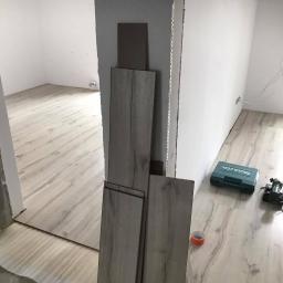 TopHome Usługi remontowo - budowlane - Montaż paneli podłogowych w jasnym odcieniu drewna, widoczne narzędzia i materiały: skrzynka narzędziowa Makita, taśma malarska, ułożone panele przy ścianie, częściowo wykończona podłoga...