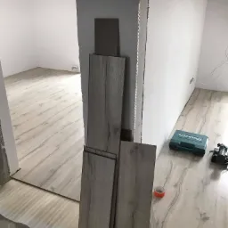 Montaż paneli podłogowych w jasnym odcieniu drewna, widoczne narzędzia i materiały: skrzynka narzędziowa Makita, taśma malarska, ułożone panele przy ścianie, częściowo wykończona podłoga...