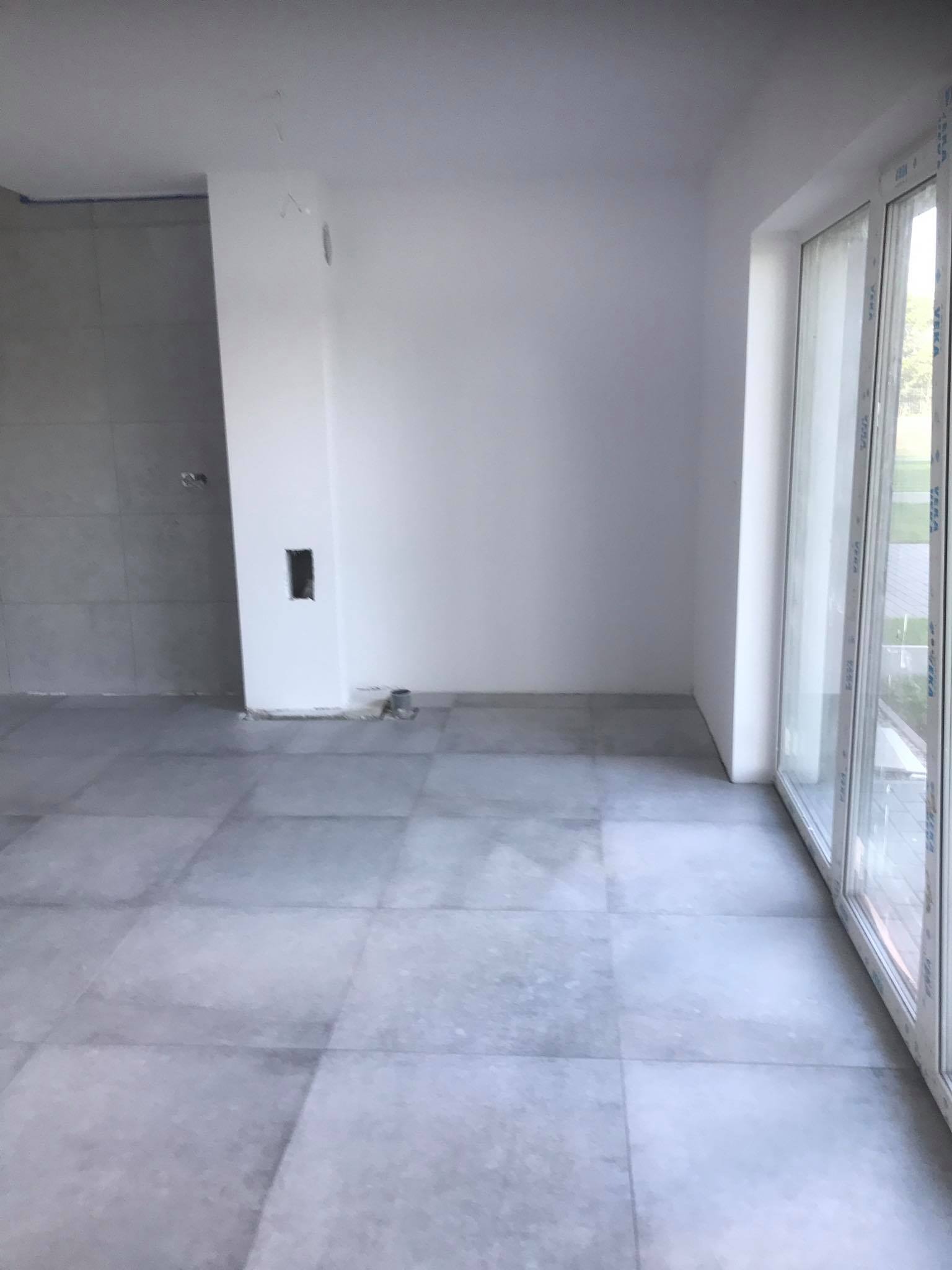 Wnętrze pokoju po remoncie z szarymi płytkami na podłodze i świeżo pomalowanymi na biało ścianami, widoczne gniazdko elektryczne w ścianie i duże okno z białymi ramami.