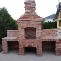 Murowany grill z cegły z wędzarnią i blatem roboczym, z metalowym rusztem wewnątrz, na tle zieleni i domu.