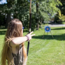 Dziewczyna z długimi włosami naciąga cięciwę łuku, celując do tarczy na zielonym trawniku, w tle drzewa i logo firmy Quatronum.