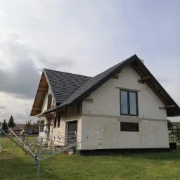 Niedokończony dom jednorodzinny z czarną dachówką, widoczne niezabezpieczone krokwie i pranie na suszarce na pierwszym planie.