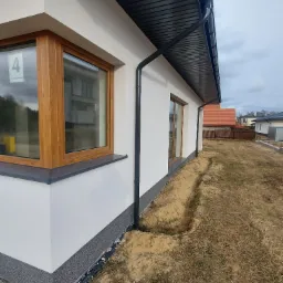 Boczny widok nowo wykończonej elewacji domu jednorodzinnego z drewnianą stolarką okienną i czarnymi rynnami, widoczny fragment terenu wok&oacute;ł budynku z wykopem pod fundament.