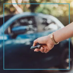 Osoba z bransoletką na nadgarstku trzyma kluczyk do ciemnego samochodu na zewnątrz, z widocznym tłem zieleni.
