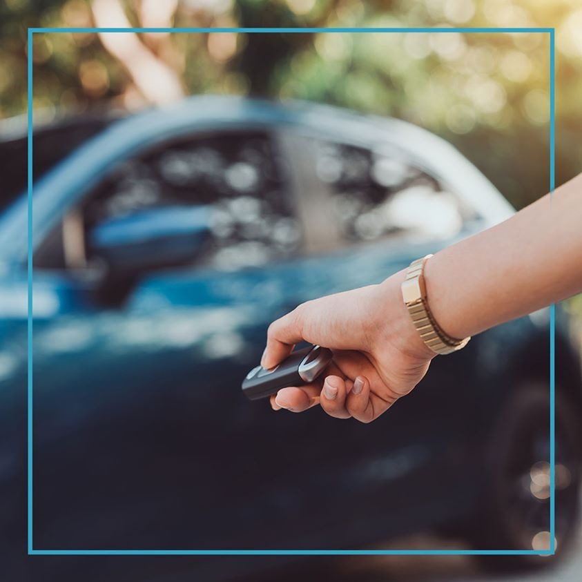 Osoba z bransoletką na nadgarstku trzyma kluczyk do ciemnego samochodu na zewnątrz, z widocznym tłem zieleni.