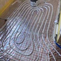 WASBUD Maciej Wąsiewski - Białe rury ogrzewania podłogowego ułożone na folii z czerwoną kratką, widoczny czarny pojemnik z klejem na górze.