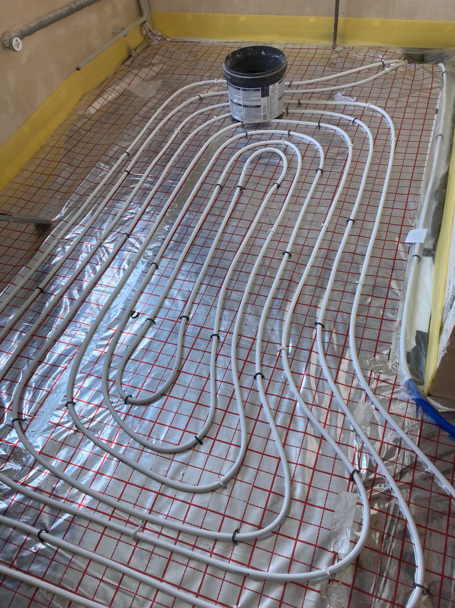 Białe rury ogrzewania podłogowego ułożone na folii z czerwoną kratką, widoczny czarny pojemnik z klejem na górze.