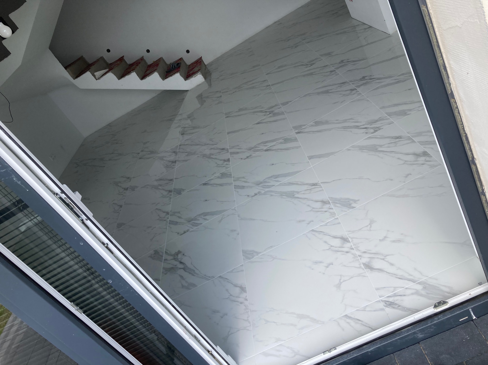 Wnętrze w trakcie wykańczania: podłoga wyłożona białymi płytkami imitującymi marmur, widoczne niedokończone elementy sufitu i fragment okna.