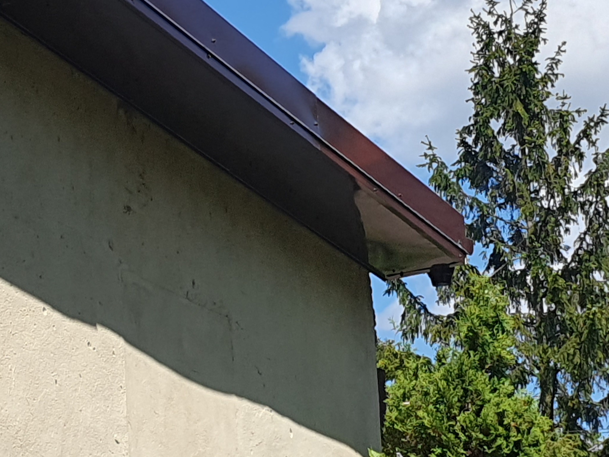 Fragment budynku z nową, brązową rynną i obróbką blacharską na tle błękitnego nieba z chmurami i zieleni drzew. Widok z dołu, podkreślający detale.