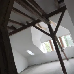 Poddasze w trakcie renowacji: surowe drewniane belki stropowe kontrastują z gładkimi, pomalowanymi na biało ścianami i sufitem, okno dachowe wpuszcza światło na szarą, betonową podłogę.