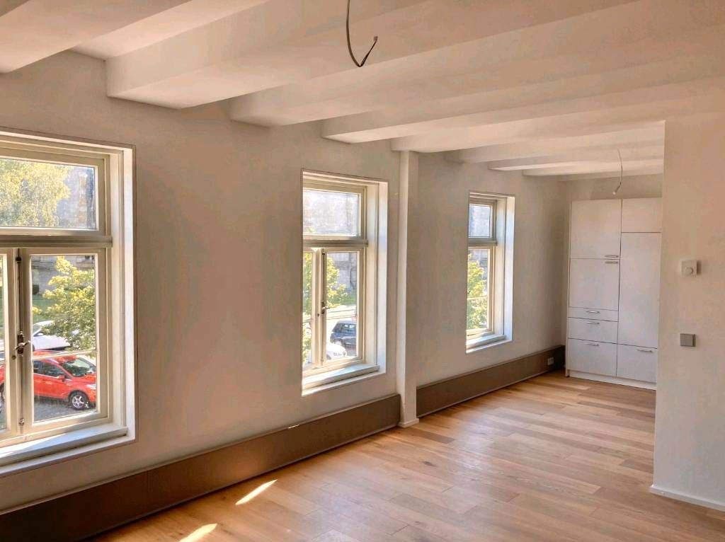 Jasne wnętrze po remoncie z odnowionymi ścianami, drewnianym parkietem i trzema oknami z widokiem na drzewa i ulicę z zaparkowanymi samochodami, w tle biała zabudowa kuchenna.