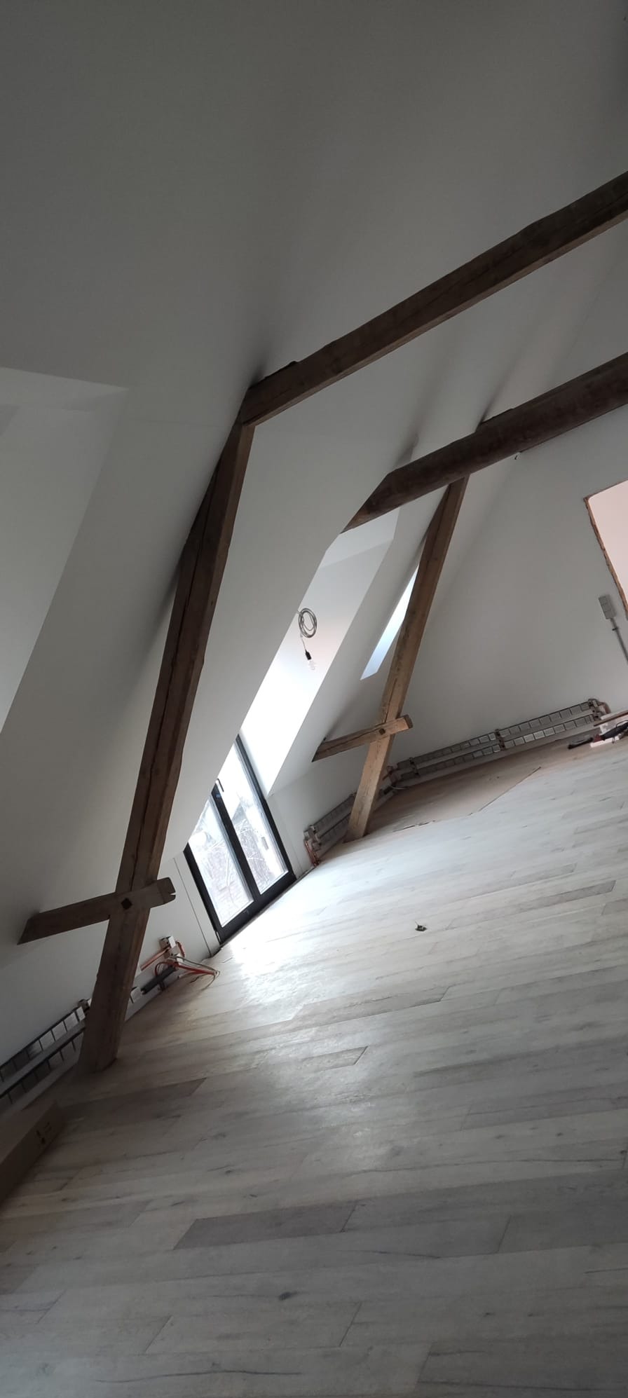 Poddasze po remoncie z drewnianymi belkami, oknem dachowym i jasną podłogą z paneli, widoczne elementy instalacji elektrycznej.