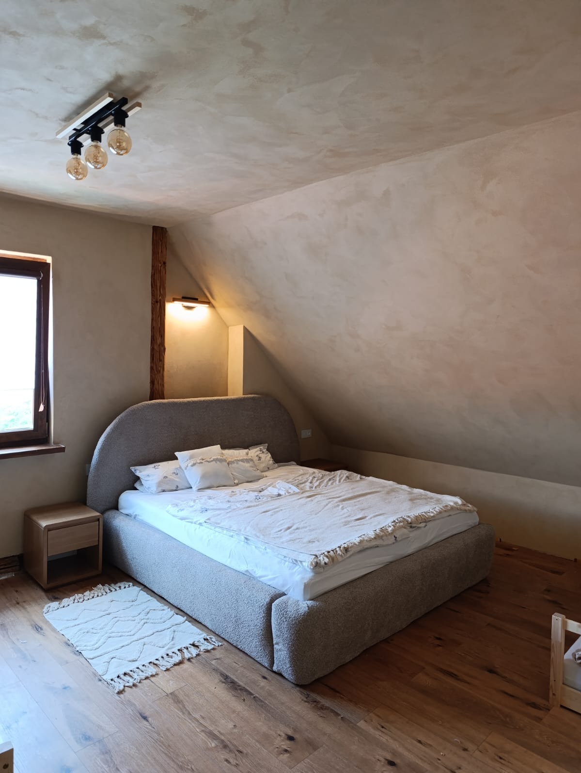 Sypialnia na poddaszu w Słopnicach z tynkiem glinianym w odcieniu Flavius na ścianach i suficie, widoczna drewniana belka i nowoczesne oświetlenie.