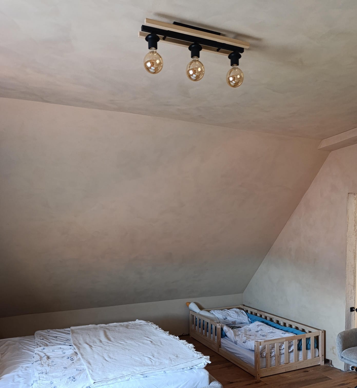 Ściana w pokoju dziecięcym pokryta tynkiem glinianym o delikatnej fakturze, oświetlona lampą sufitową z trzema żarówkami w stylu retro, poniżej widoczne dwa łóżka dziecięce.