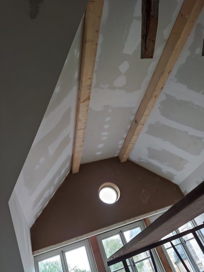 Skośny sufit poddasza z zabudową z płyt gipsowo-kartonowych, drewnianymi belkami konstrukcyjnymi i okrągłym oknem w szczycie ściany, widoczne ślady szpachlowania.