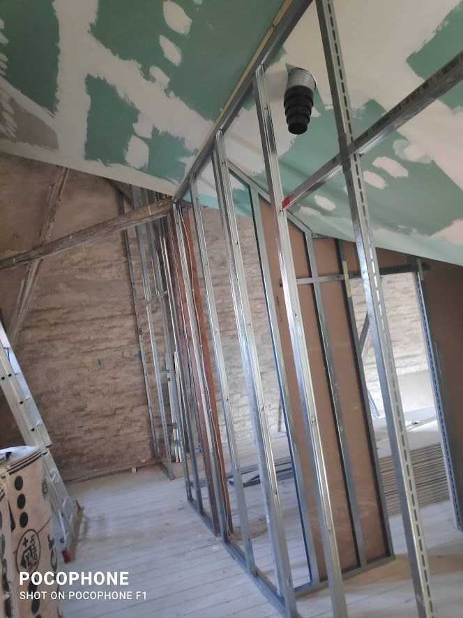 Stelaż metalowy do ścianki działowej z płyt gipsowo-kartonowych w trakcie montażu, widoczny fragment starej ściany z cegły, sufit częściowo pokryty gładzią gipsową, rura wentylacyjna w suficie.