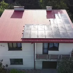 Dom jednorodzinny z częściowo zamontowanymi panelami słonecznymi na czerwonym dachu z blachy trapezowej, widok z góry.